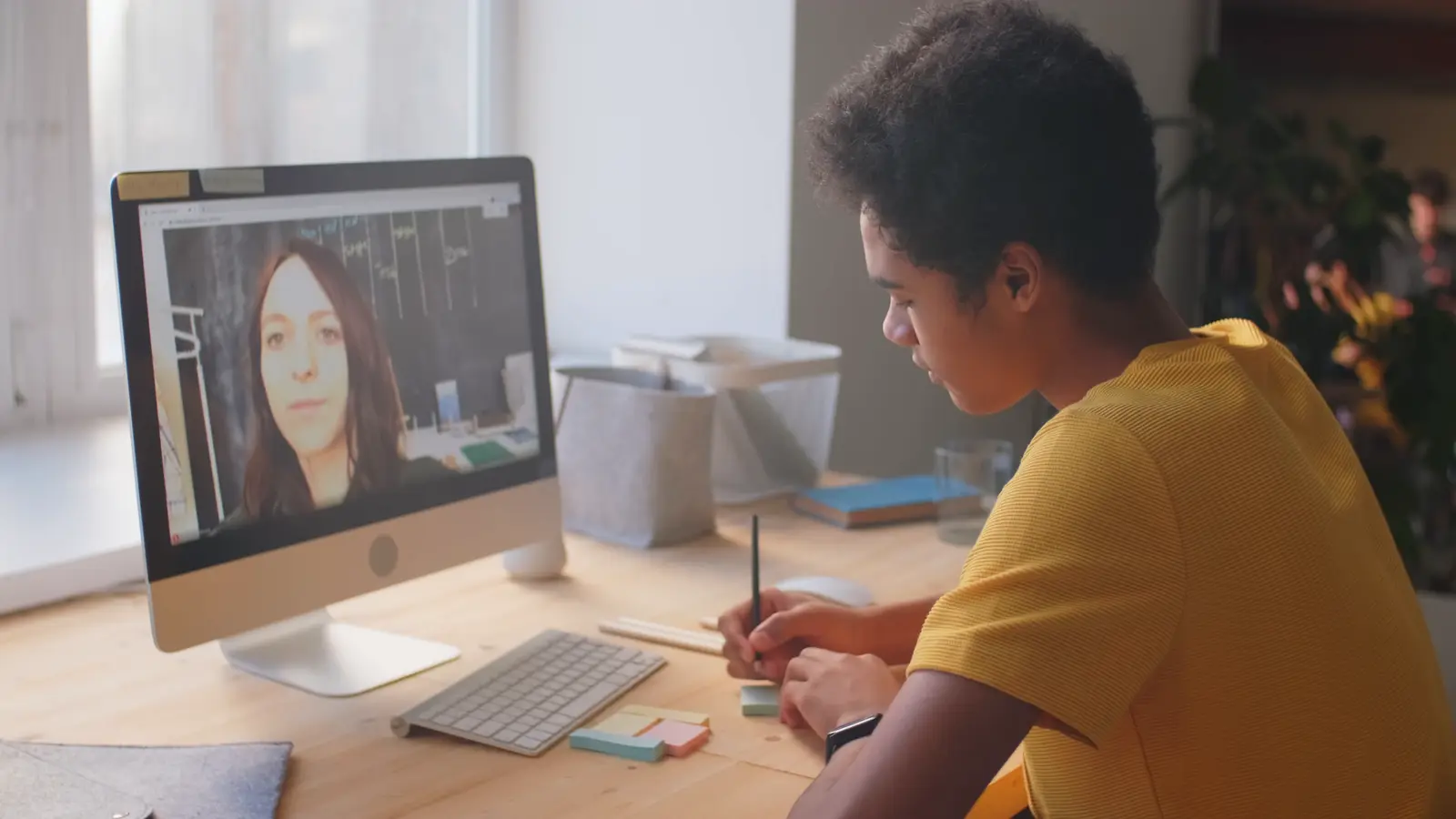 A boy in a yellow T-shirt writing notes during a video lesson