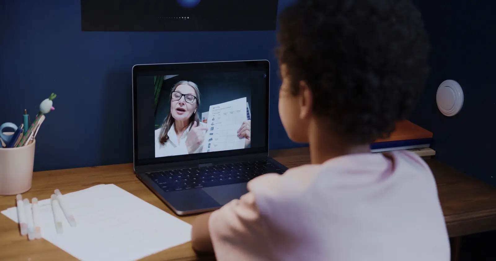 A child at a desk on a video lesson, the teacher holding up a page on screen