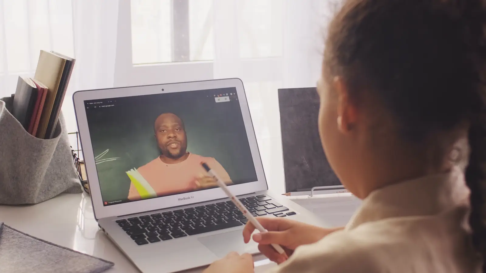 A girl with a pencil watching her teacher explain something on a laptop video call