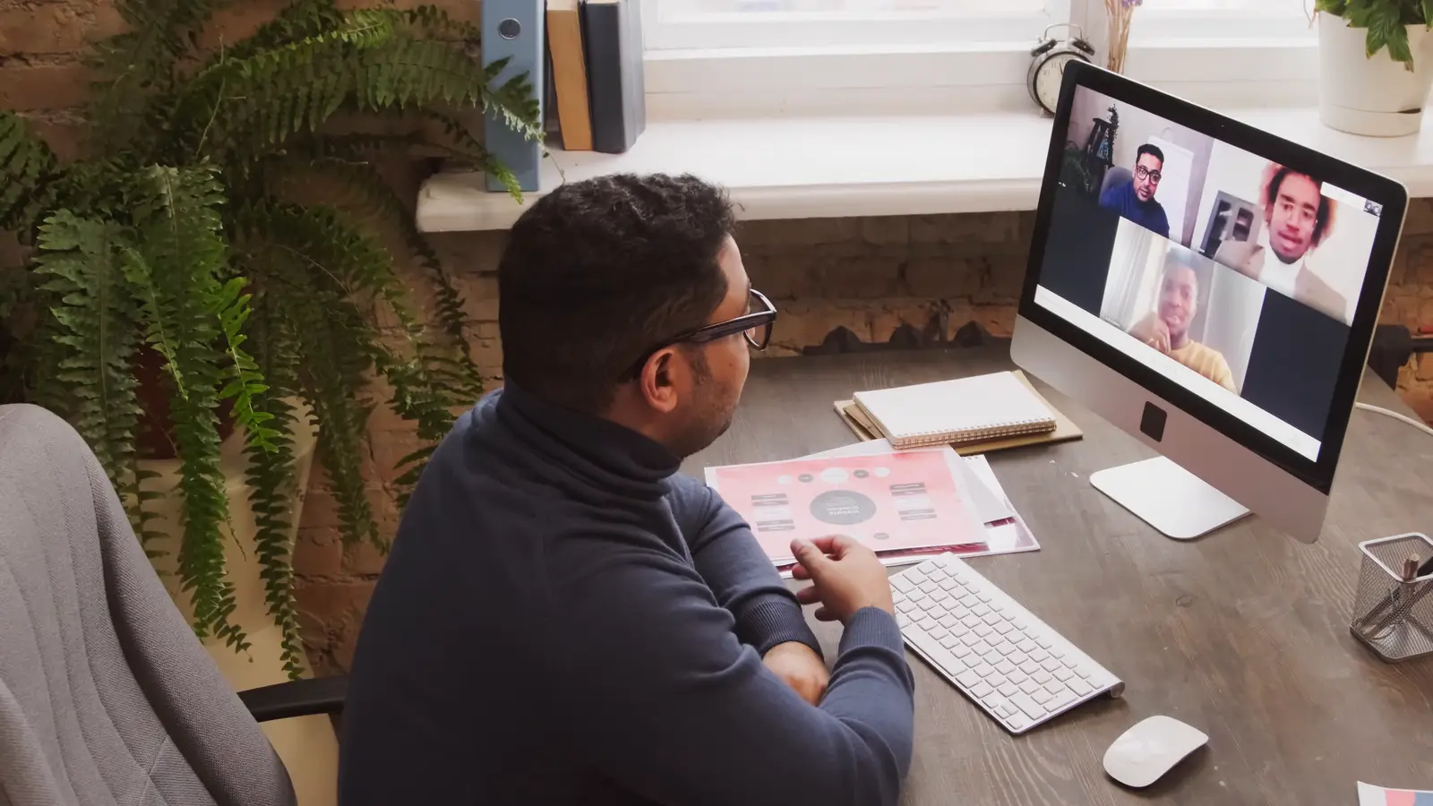 A man at a desk by a window on a video call with three people