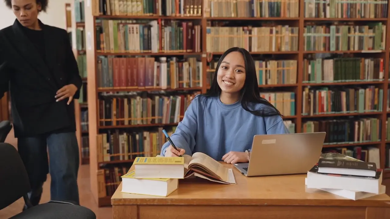 A student smiling at a library table with open books and a laptop