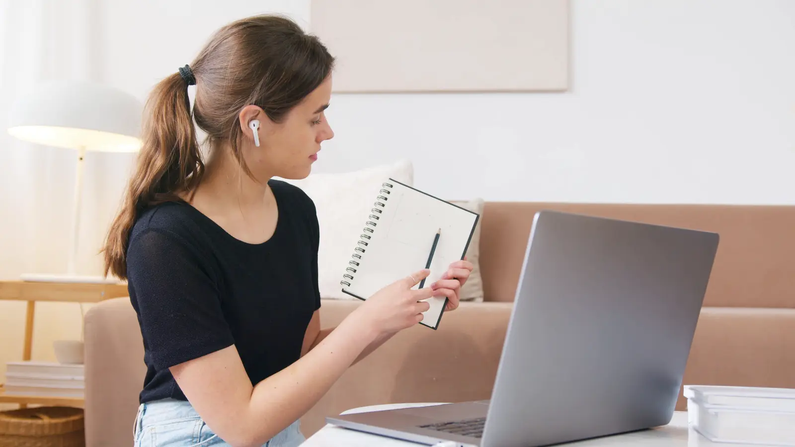 A woman with earphones holding up a notebook to her laptop on a video call