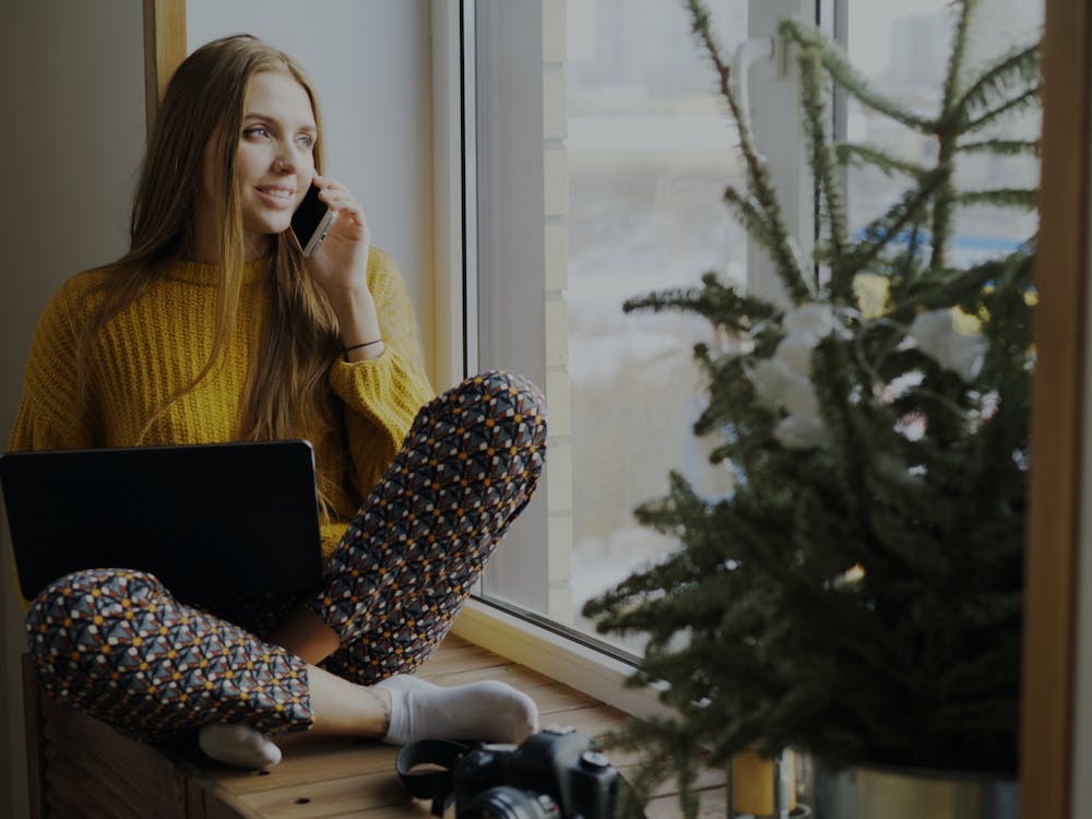 A woman sitting by a window with a laptop, talking on her phone and smiling.
