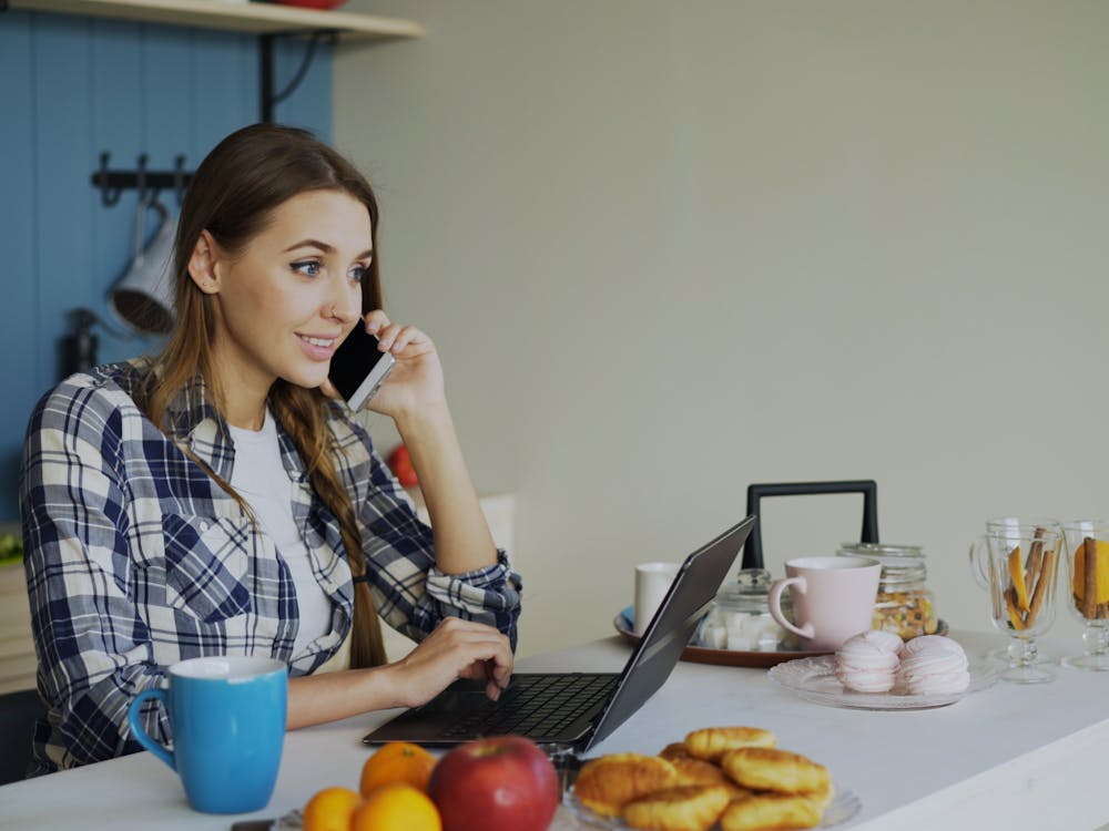 A woman at her kitchen counter on the phone with a laptop open in front of her.