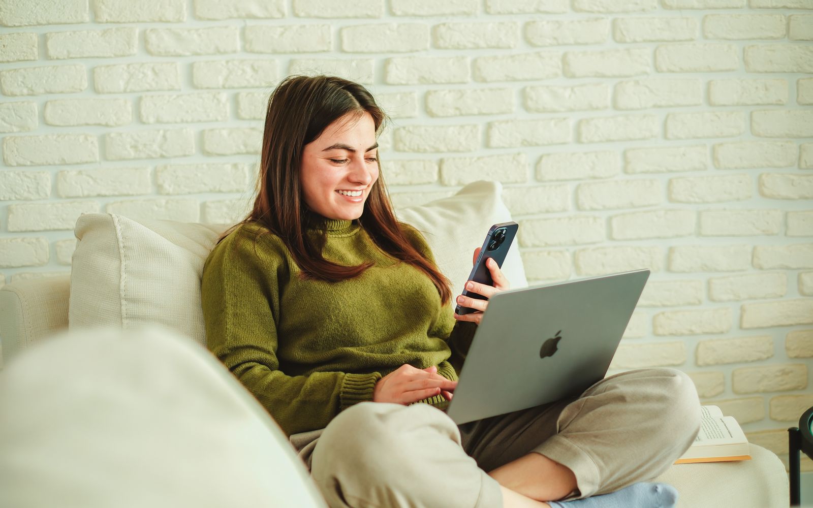 A woman relaxing on her sofa with a laptop on her lap, smiling at her phone.