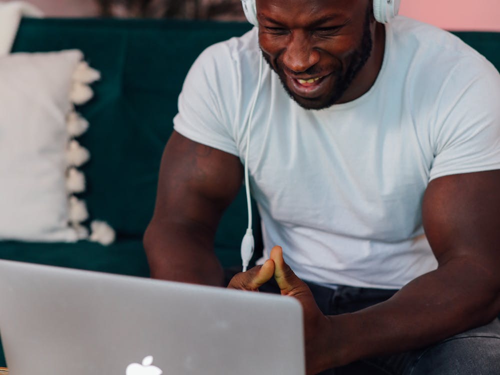 A man in a white t-shirt and headphones laughing during a video call at his laptop.