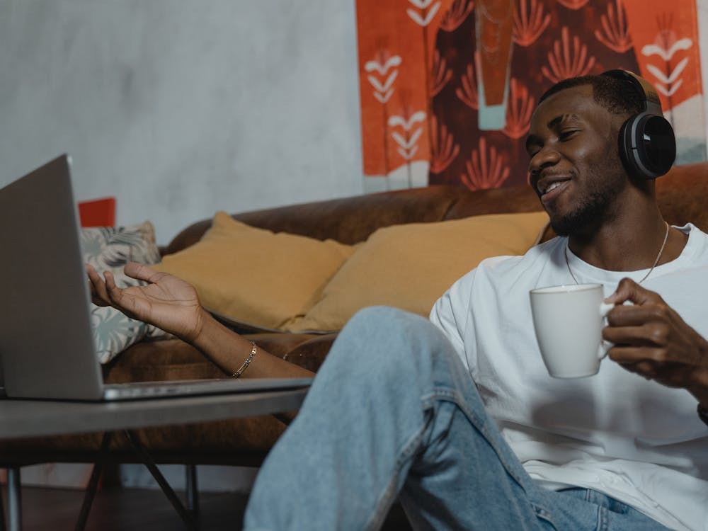 A man with headphones and a mug gesturing at his laptop on the sofa.