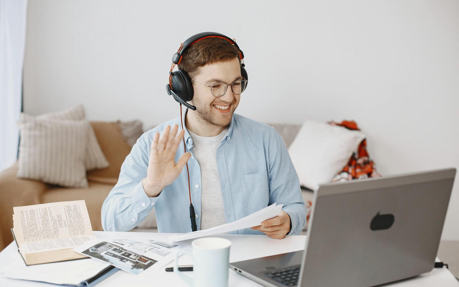 A man in a blue shirt and headphones smiling and gesturing during a video conversation at his laptop.