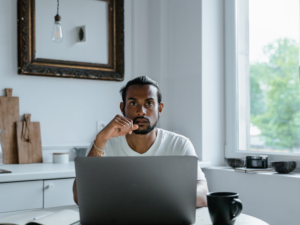 A man at his kitchen table with a laptop, resting his chin on his hand.