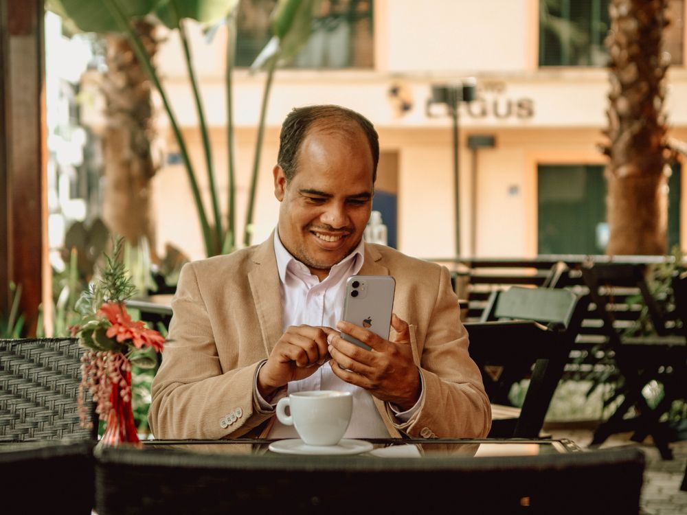 A man in a blazer smiling at his phone at an outdoor café table.