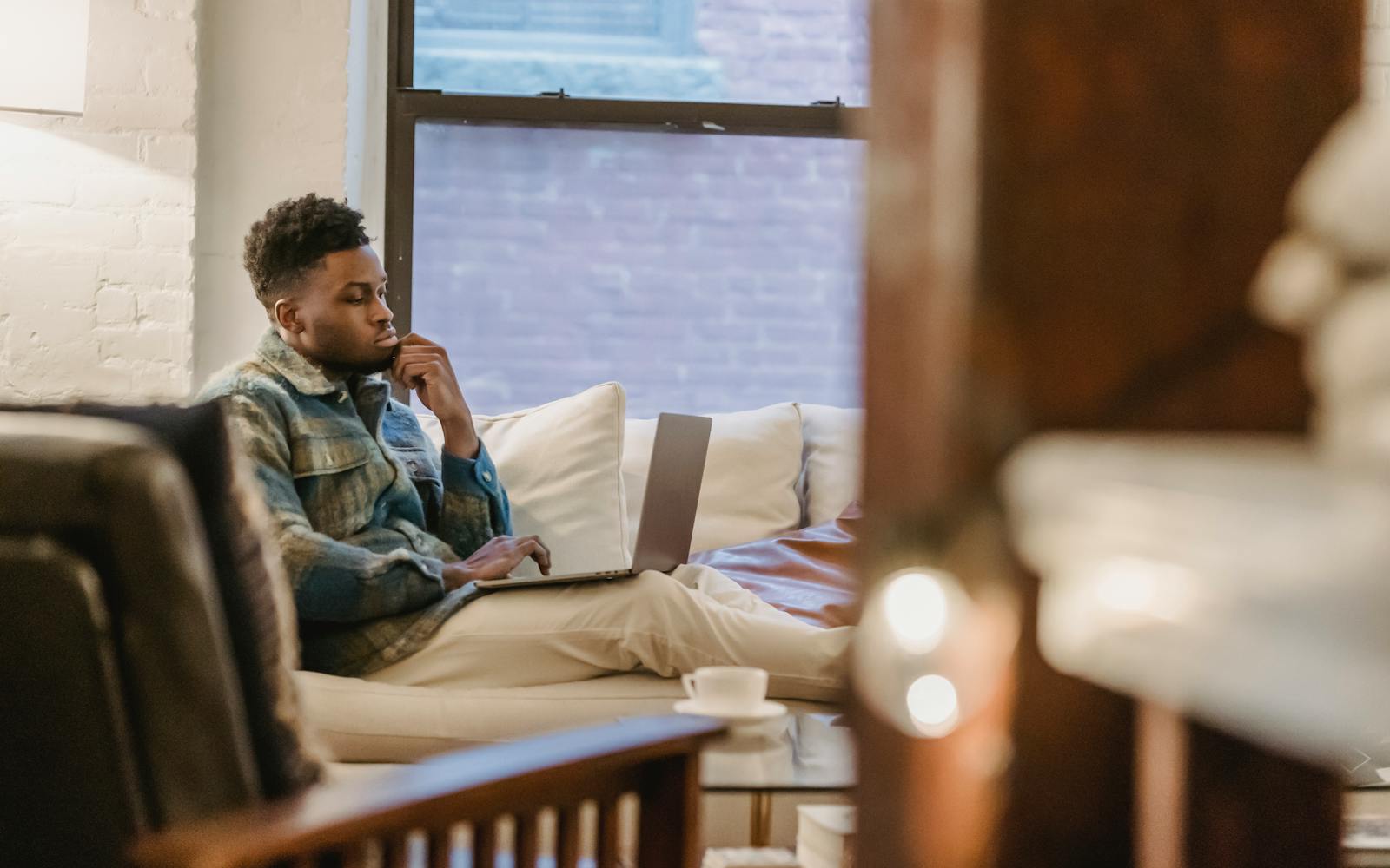 A man sitting on his sofa by the window with a laptop, thinking.