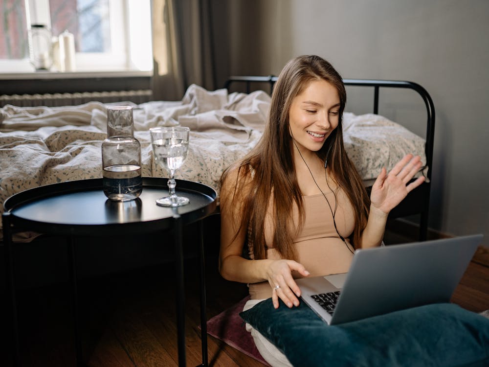 A woman sitting at home, smiling and waving at her laptop during a relaxed video conversation.