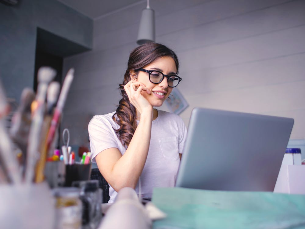 A woman in glasses smiling at her laptop at a home desk.