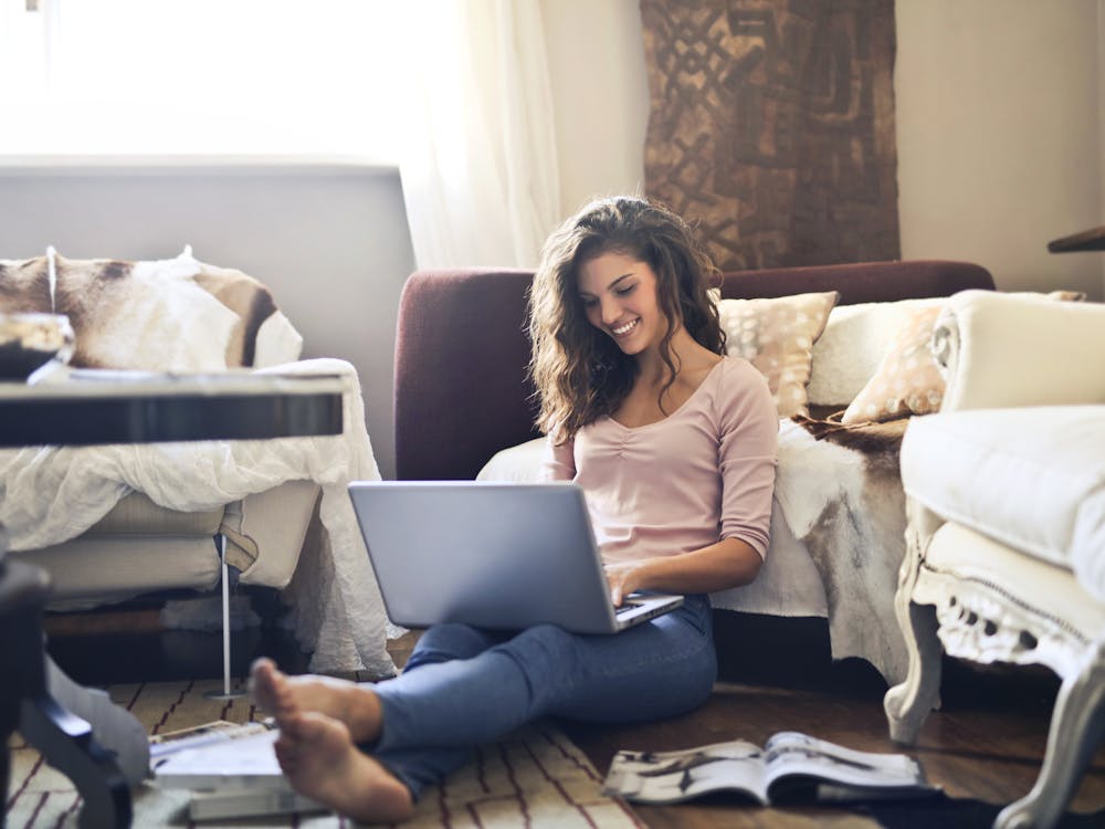 A woman sitting on the floor against her sofa, smiling at her laptop.