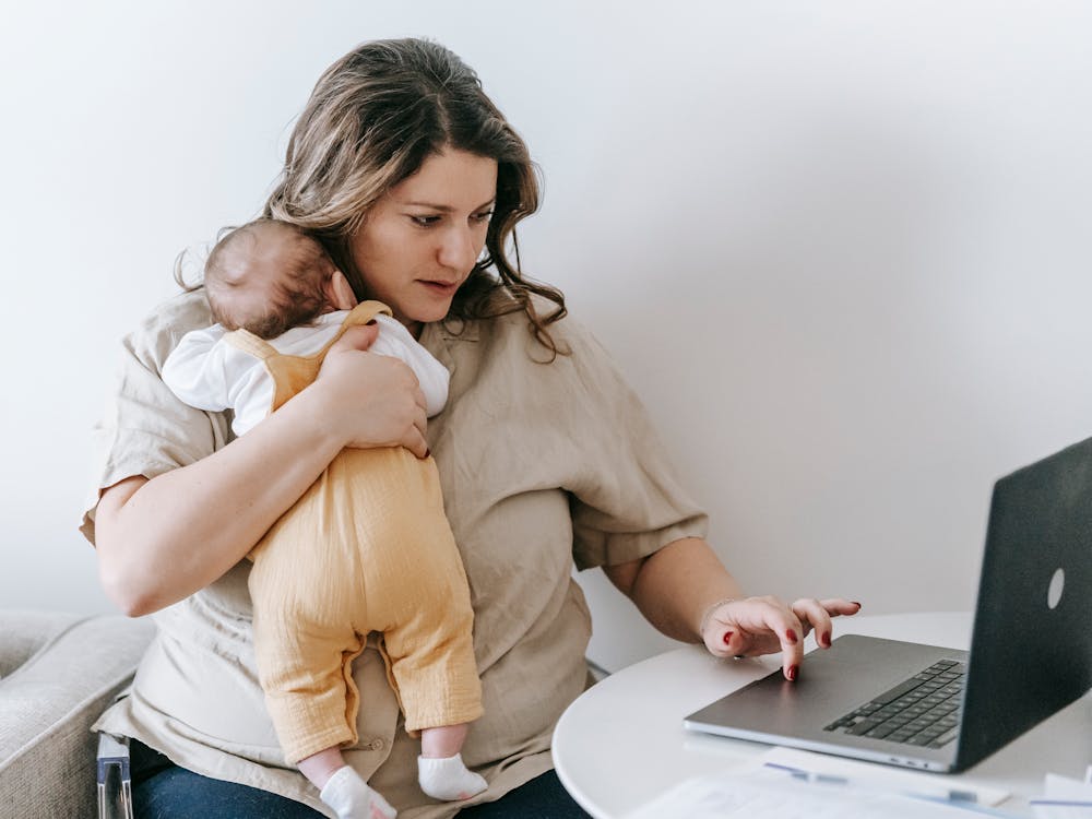 A mother holding her baby on one arm while using a laptop at a table.