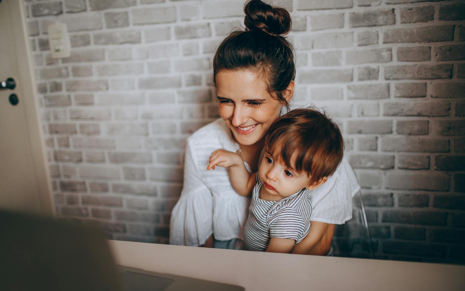 A mother holding her toddler, smiling at a laptop at home.