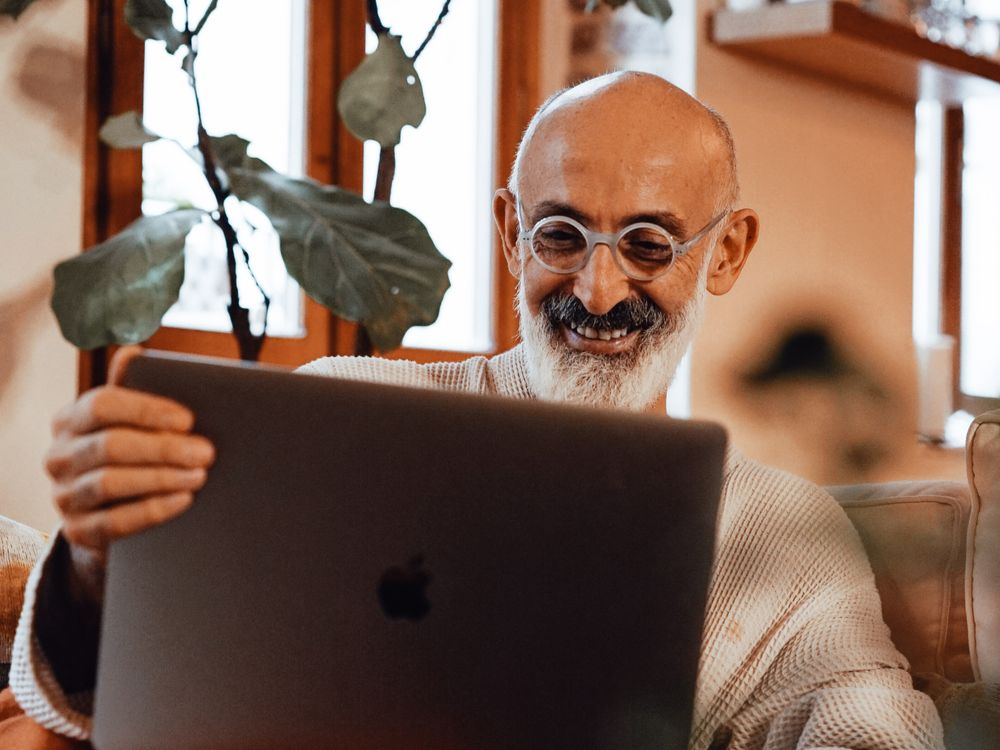 An older man with glasses smiling warmly at his laptop at home.