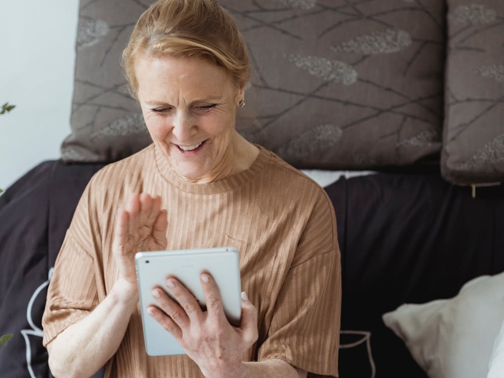 An older woman smiling and waving at her tablet during a video call.