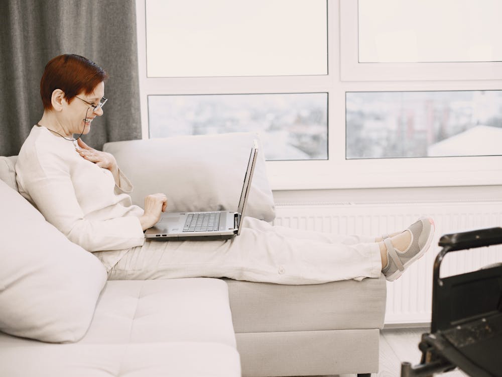 An older woman relaxing on her sofa with a laptop, smiling.