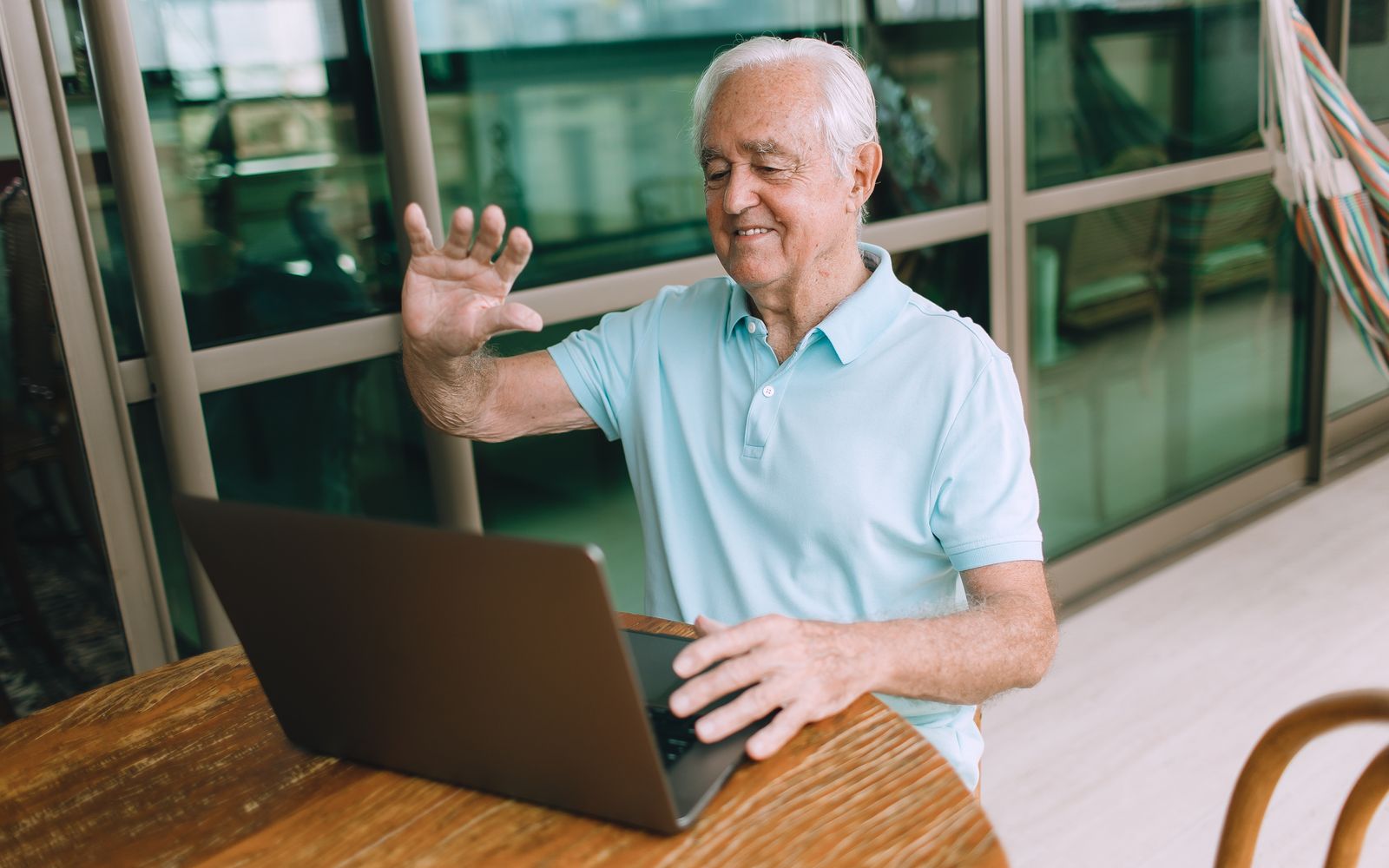 An older man in a light blue shirt smiling and waving at his laptop during a video call on a balcony.
