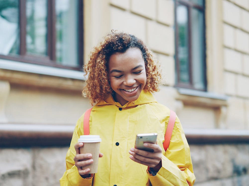 A young woman in a yellow raincoat smiling down at her phone on a city street.