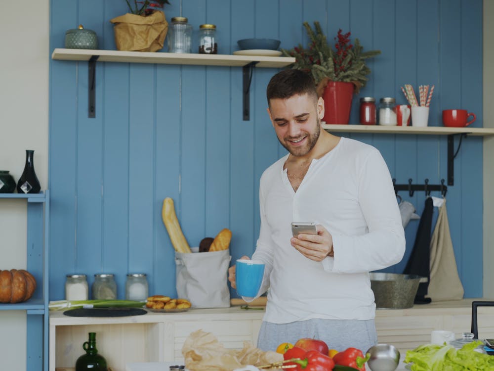 A man smiling at his phone with a coffee in a bright kitchen.