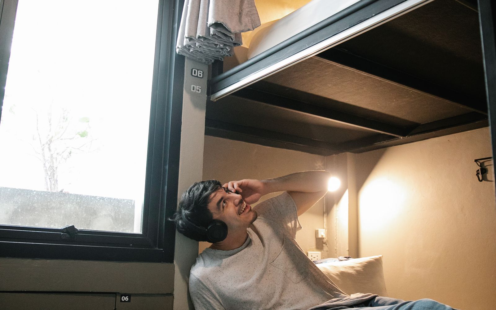 A young traveller in headphones smiling against a bright window in a hostel.