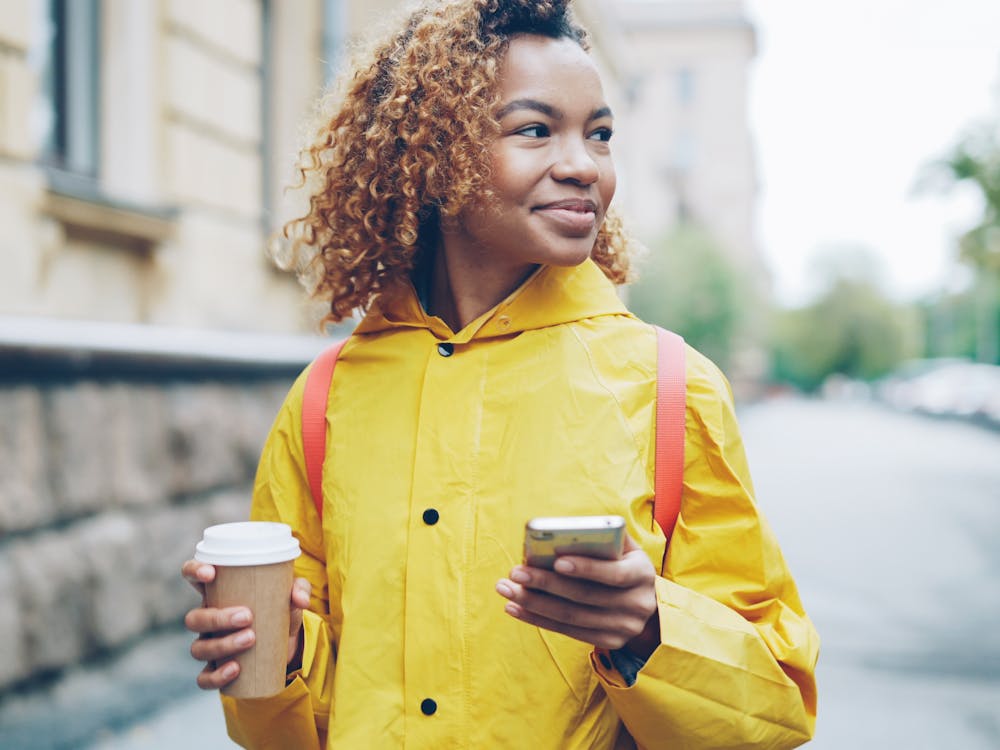 A woman outdoors with a coffee, smiling at her phone.