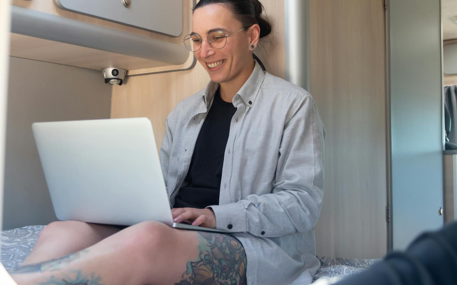 A woman sitting on the bed inside her camper van, smiling at a laptop on her knees.